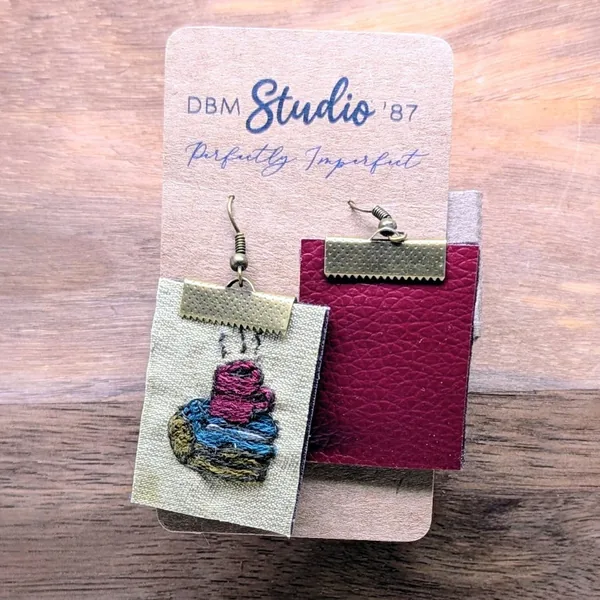 A pair of embroidered earrings with a coffee cup on a stack of books pictured on them. With a maroon back.